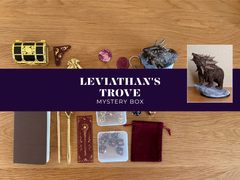 Leviathan's Trove Mystery Box contents arranged on a wooden tabletop, including a small gold-and-black treasure chest, crystals, a dark metallic figurine, dice, a notebook, rods, a velvet pouch, and a central purple banner reading 'LEVIATHAN'S TROVE MYSTERY BOX'.