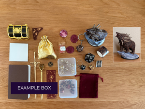 A tabletop flat lay of a themed TTRPG fantasy adventurer box set on a light wood background, featuring a gold and black miniature treasure chest, a gold drawstring pouch, a gold sword, a brown journal, dice, and a bear figurine.