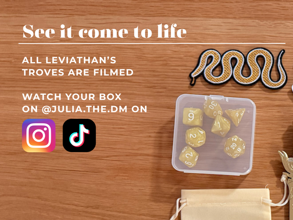 A promotional flat-lay on a wooden surface showing a clear plastic box containing yellow polyhedral dice, a small cream drawstring pouch, a coiled snake patch, and social media text reading 'See it come to life' with Instagram and TikTok icons.